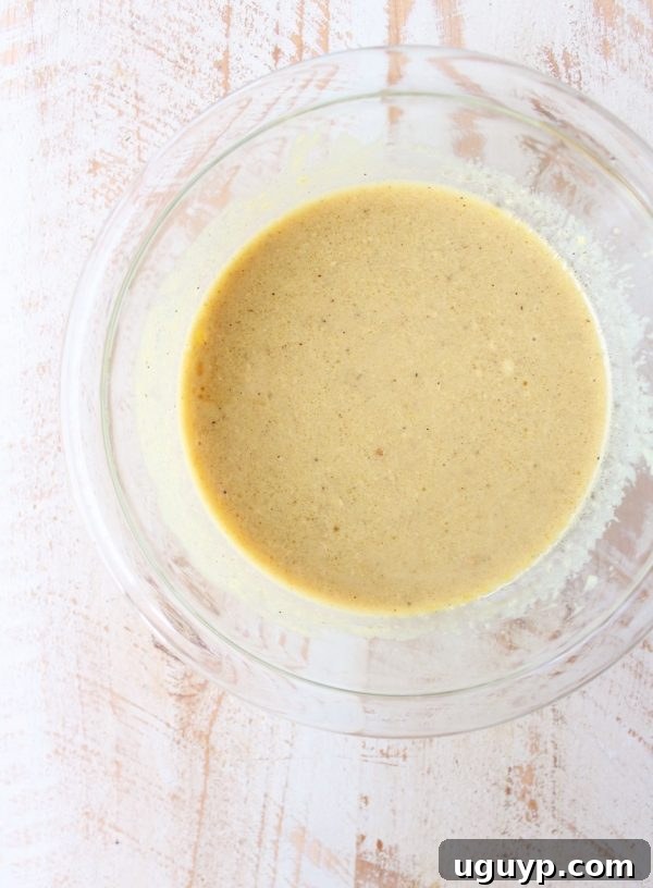 A bowl of honey dijon dressing next to a whisk, illustrating the first step of preparing the salad dressing, with fresh garlic cloves and lemon wedges in the background.