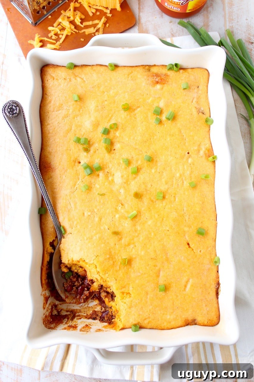 A bubbling casserole dish filled with Buffalo Cheddar Cornbread Chili Pie, ready to be served, with a large spoon scooping out a portion.