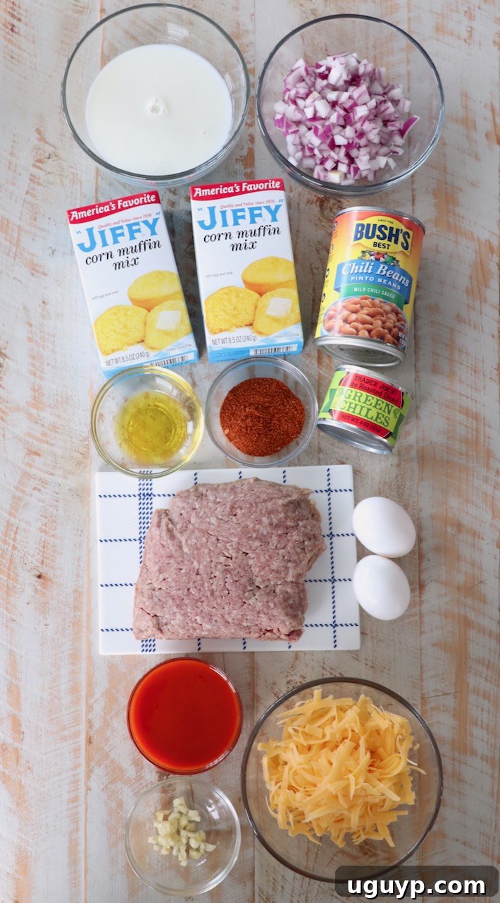 Ingredients for Buffalo Cheddar Cornbread Chili Pie laid out on a white wooden board, including fresh vegetables, ground beef, and various canned goods.