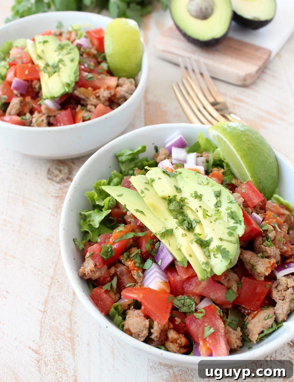 Two white bowls, artfully arranged on a rustic wooden surface, brimming with Turkey Taco Salad. Each bowl contains a base of fresh lettuce, generously topped with savory taco-seasoned ground turkey, colorful peppers, onions, tomatoes, and slices of creamy avocado.