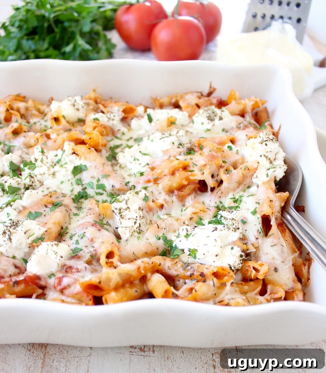 Close-up of baked ziti in a casserole dish, topped with generous dollops of creamy ricotta cheese and a sprinkle of fresh green herbs, promising a rich and flavorful bite.