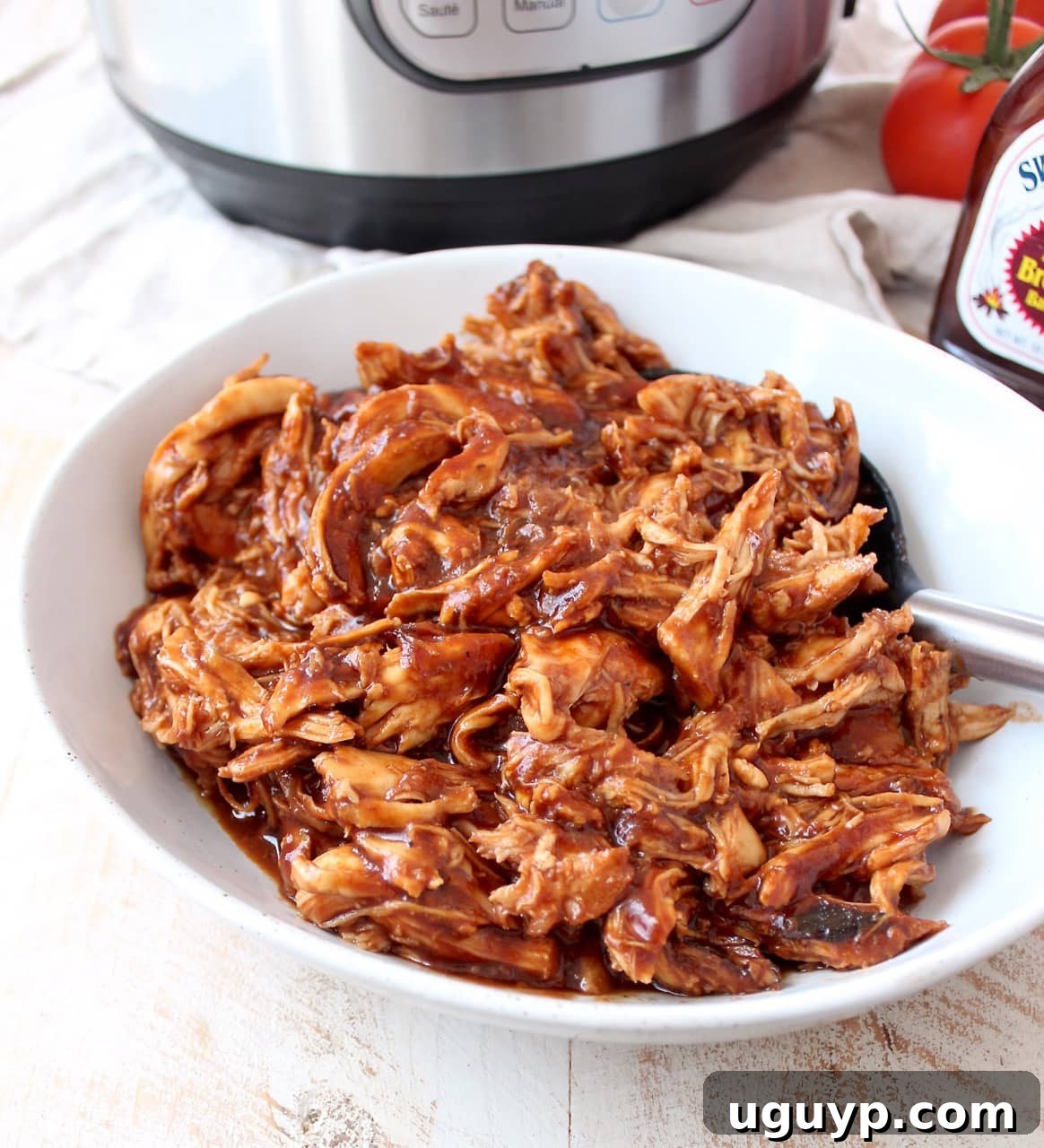 Close-up of shredded BBQ chicken in a white bowl with a serving spoon, garnished with fresh herbs.