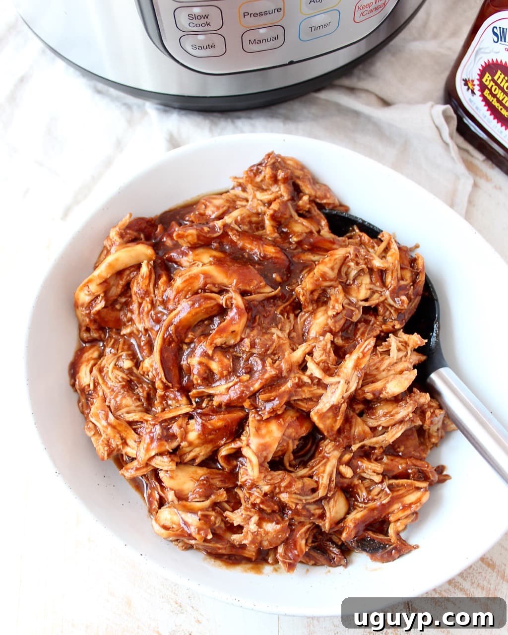 A large white bowl filled with shredded BBQ chicken, garnished with fresh cilantro and a serving spoon.