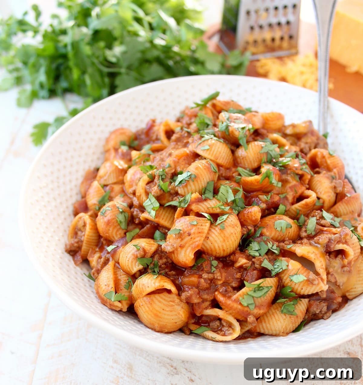 A large bowl filled with creamy enchilada pasta, showcasing perfectly cooked pasta shells coated in a rich, meaty red sauce and topped with melted cheddar cheese. A serving spoon is dug in, ready for a delicious helping.