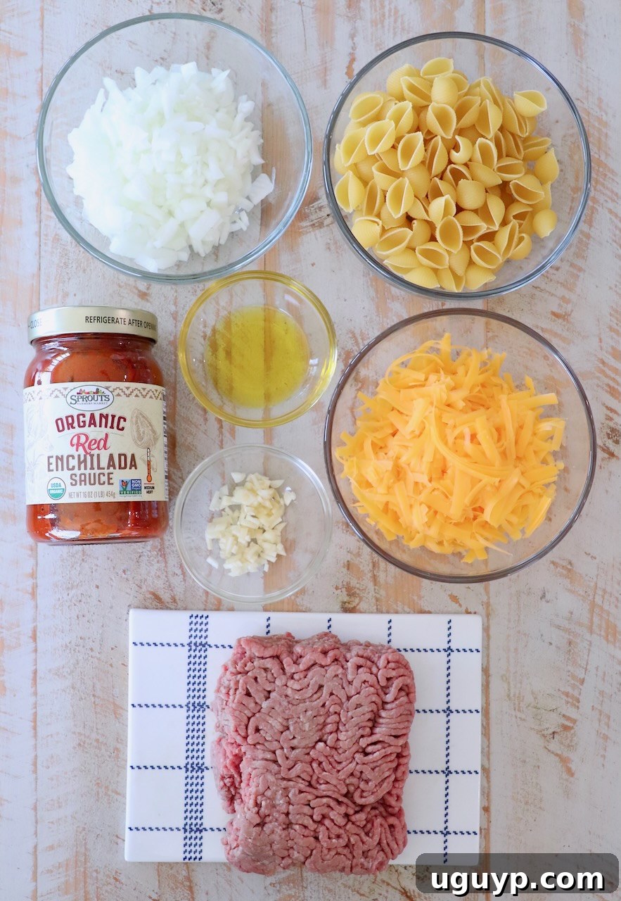 Seven essential ingredients for Enchilada Pasta neatly arranged on a white wooden board: uncooked pasta shells, a bottle of red enchilada sauce, shredded cheddar cheese, ground beef, onion, garlic, and olive oil.