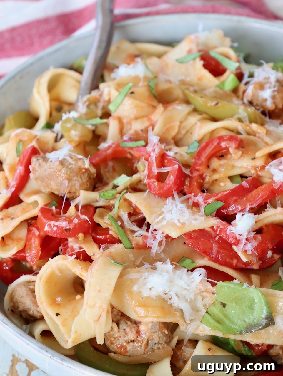 A bowl of cooked Italian Drunken Noodles tossed with colorful peppers, sliced Italian sausage, and fresh basil leaves, with a fork ready to serve.