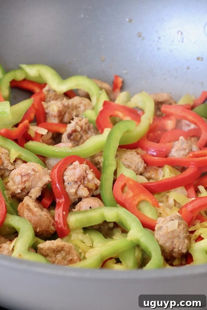 Sliced bell peppers and spicy Italian sausage browning in a large pan with the sautéed onions and pepperoncinis.