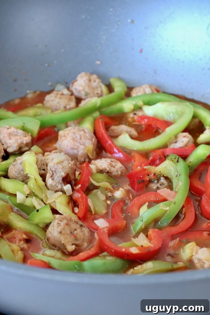 Sliced bell peppers and Italian sausage simmering in a rich broth and tomato sauce in a large pan.