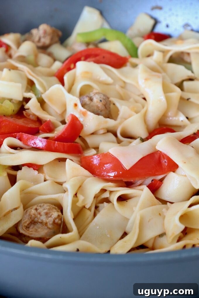 Cooked wide egg noodles tossed with the bell peppers, sausage, and sauce in a large pan, ready for the final touches.