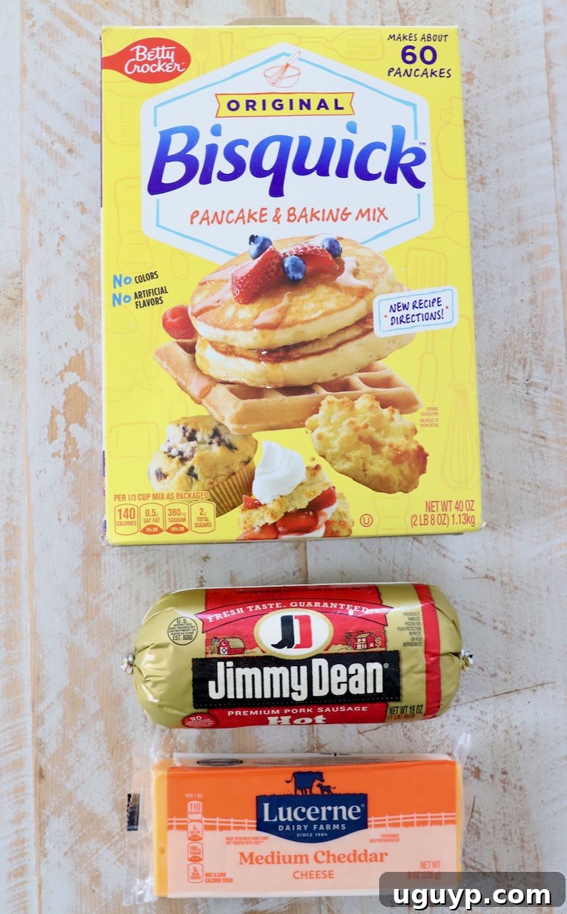 Ingredients for Bisquick Sausage Balls: a block of cheddar cheese, a roll of ground sausage, and a box of Bisquick mix laid out on a white wooden board.