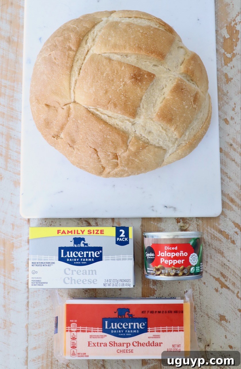 A visually appealing shot of the raw ingredients: a round loaf of sourdough bread on a marble board, accompanied by a block of Lucerne cream cheese, a wedge of extra sharp cheddar, and a small bowl of diced jalapeño peppers, all elegantly arranged on a rustic wooden table.