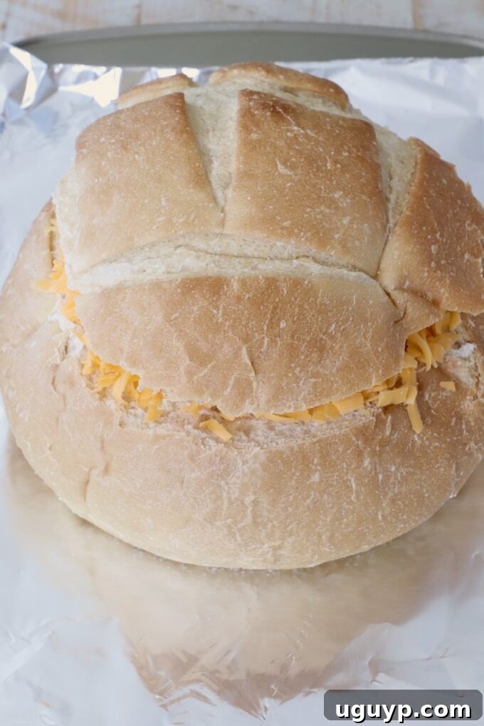 A round bread loaf with its top sliced off and the interior carefully hollowed, now filled with shredded cheese, resting on a sheet of aluminum foil on a baking sheet, ready for the next step.