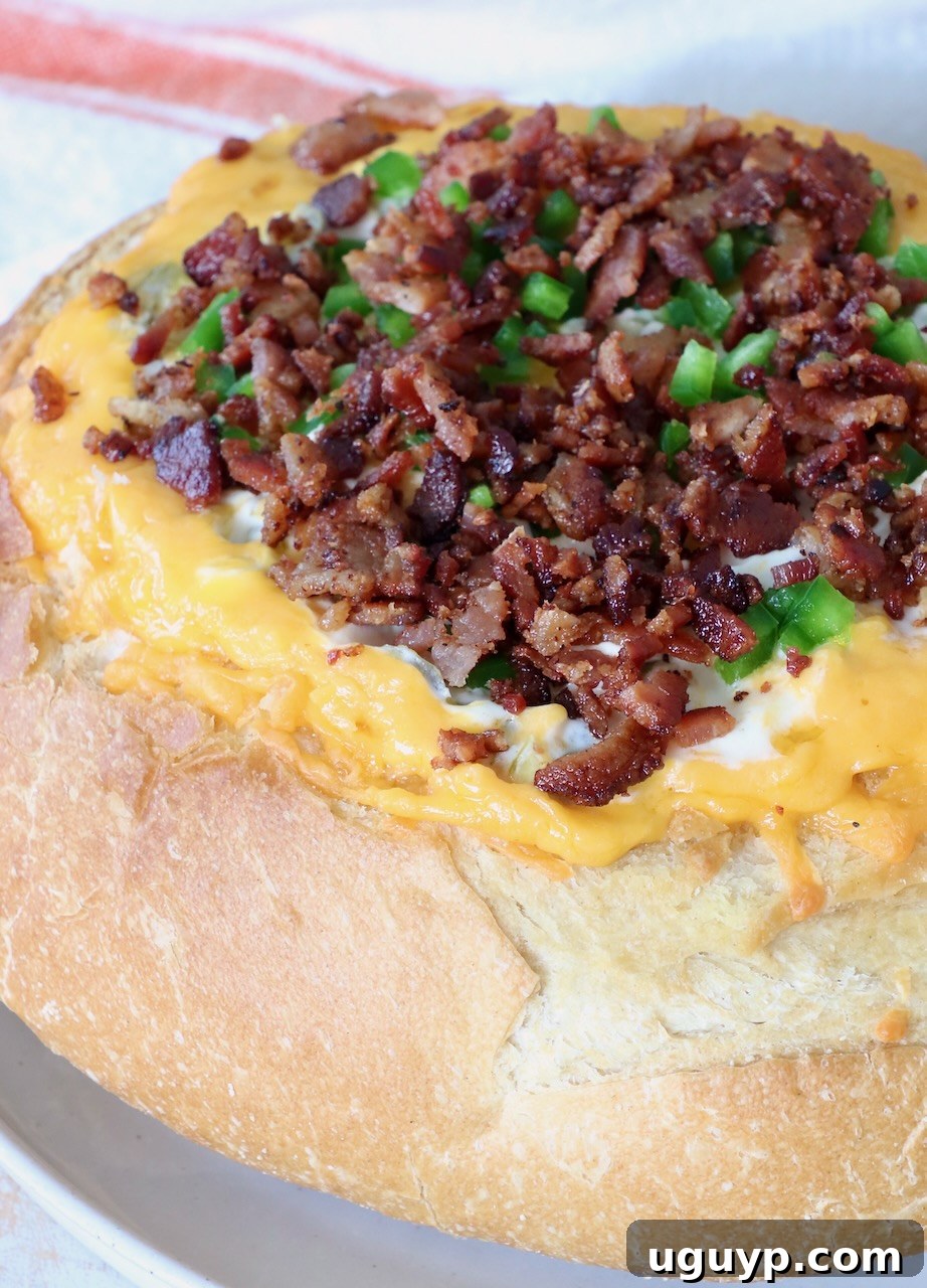 A perfectly baked, round bread loaf, hollowed and generously filled with melted, golden cheese, generously topped with crispy crumbled bacon bits and vibrant chopped green peppers, ready to be served as an irresistible appetizer.