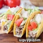 Ground beef tacos in crispy shells with diced tomatoes and shredded lettuce on wood cutting board with whole tomatoes in back