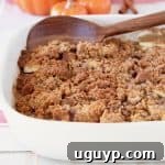 Apple crumble in a white baking dish with a wooden spoon, surrounded by ceramic orange pumpkins and cinnamon sticks in the background.