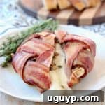 Bacon Wrapped Brie with melted brie spilling out onto plate with fresh herbs on plate and sliced baguette on wood cutting board in background