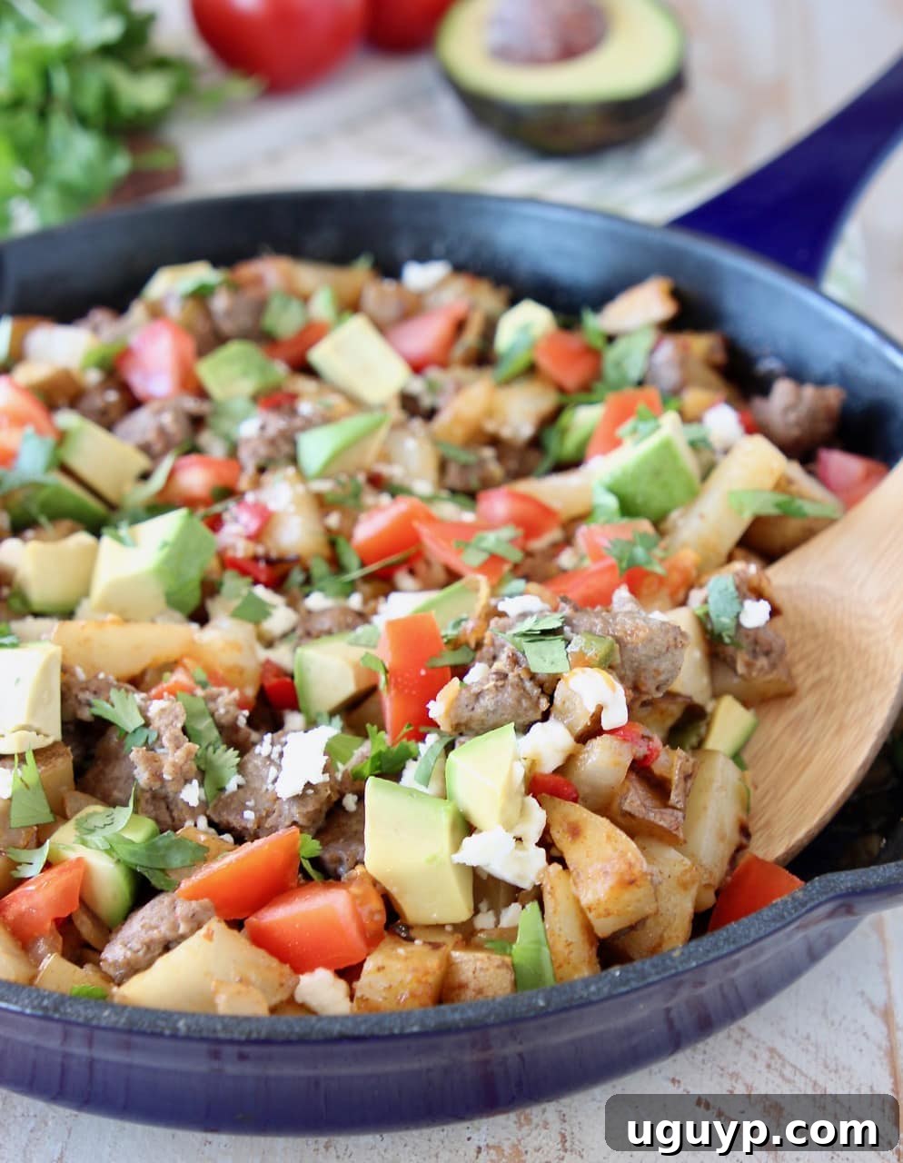 Vibrant breakfast hash in a blue cast iron skillet, generously topped with diced avocado, fresh tomatoes, crumbled turkey sausage, and perfectly cooked potatoes. A wooden spoon rests in the skillet, with fresh cilantro scattered in the background, hinting at the dish's fresh flavors.