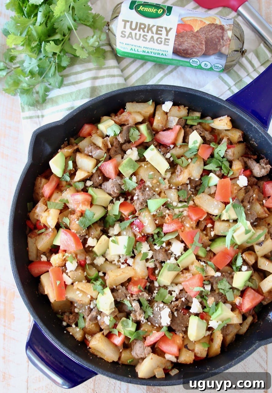 Close-up of a blue cast iron skillet brimming with a delicious breakfast hash. The dish is loaded with perfectly diced potatoes, savory turkey sausage crumbles, creamy avocado, fresh tomatoes, and delicate queso fresco. A sprinkle of fresh cilantro adds a touch of brightness, with a package of Jennie-O turkey sausage subtly placed in the background.