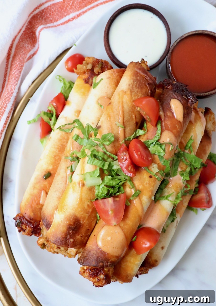 cooked taquitos stacked up on a plate topped with buffalo sauce, tomatoes, and shredded lettuce