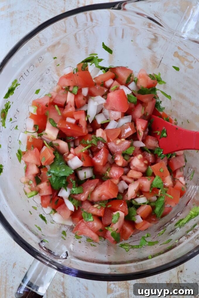 Freshly mixed Pico de Gallo in a glass bowl with a red spatula, ready to be served or chilled.