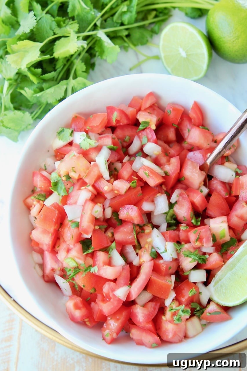 Overhead shot of fresh Pico de Gallo in a white bowl, accompanied by fresh lime wedges and a spoon, with cilantro on the side.