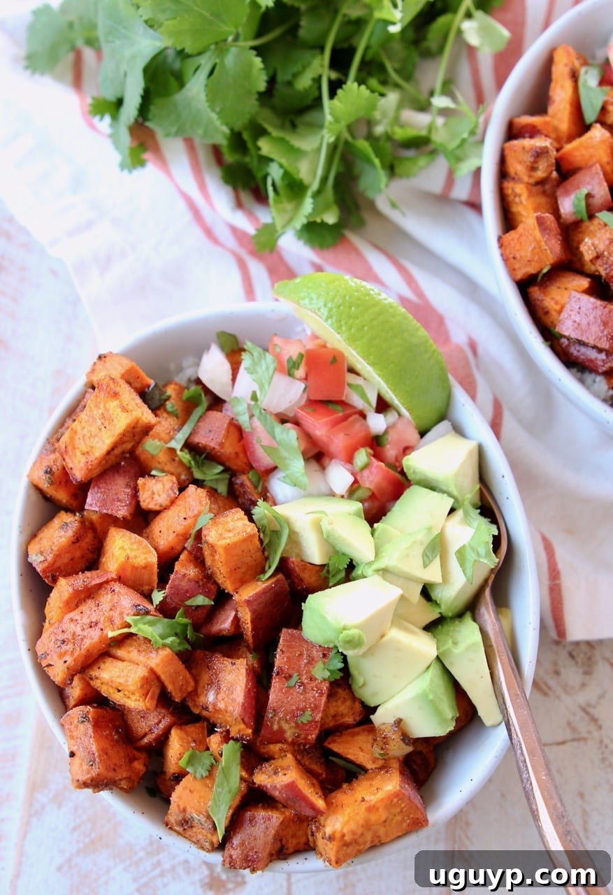 Vibrant roasted diced sweet potatoes in a bowl, generously topped with fresh diced avocado, zesty pico de gallo, a bright lime wedge, and accompanied by a copper fork. A stylish orange and white striped towel is visible beneath, with fresh cilantro adding a touch of green in the background, creating an inviting culinary scene.
