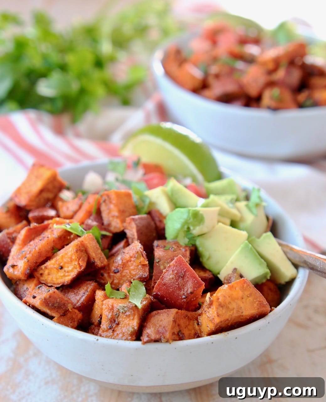 Close-up overhead view of a colorful quinoa bowl featuring golden roasted sweet potato cubes, creamy diced avocado, and fresh pico de gallo, with a bright lime wedge and a fork resting beside it. A textured orange and white striped towel provides a cozy backdrop, alongside hints of fresh cilantro.