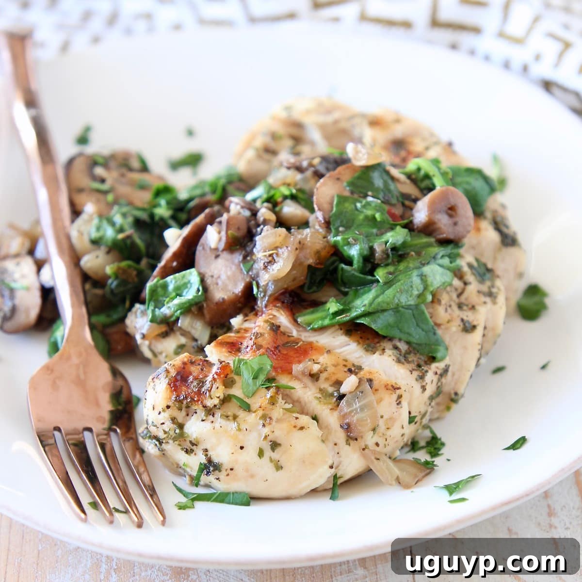 A white plate featuring perfectly sliced spinach mushroom chicken, accompanied by a gleaming copper fork, all resting on a elegant gold and white patterned napkin.