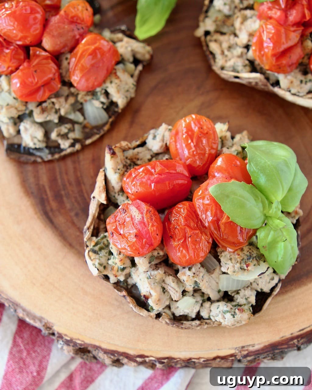Overhead view of perfectly baked Italian Stuffed Portobello Mushrooms, featuring a savory ground turkey and sweet cherry tomato filling, garnished with fresh basil leaves, presented on a round, raw-edge wooden cutting board, set against a cozy red and white striped towel.
