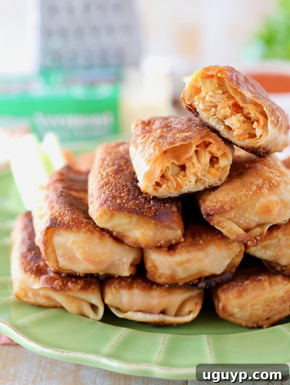 Crispy pan-fried Buffalo chicken egg rolls piled high on a vibrant green plate, with one egg roll cut in half to showcase the melted cheddar cheese and shredded chicken filling. A cheese grater and block of cheddar are blurred in the background, hinting at the fresh ingredients.
