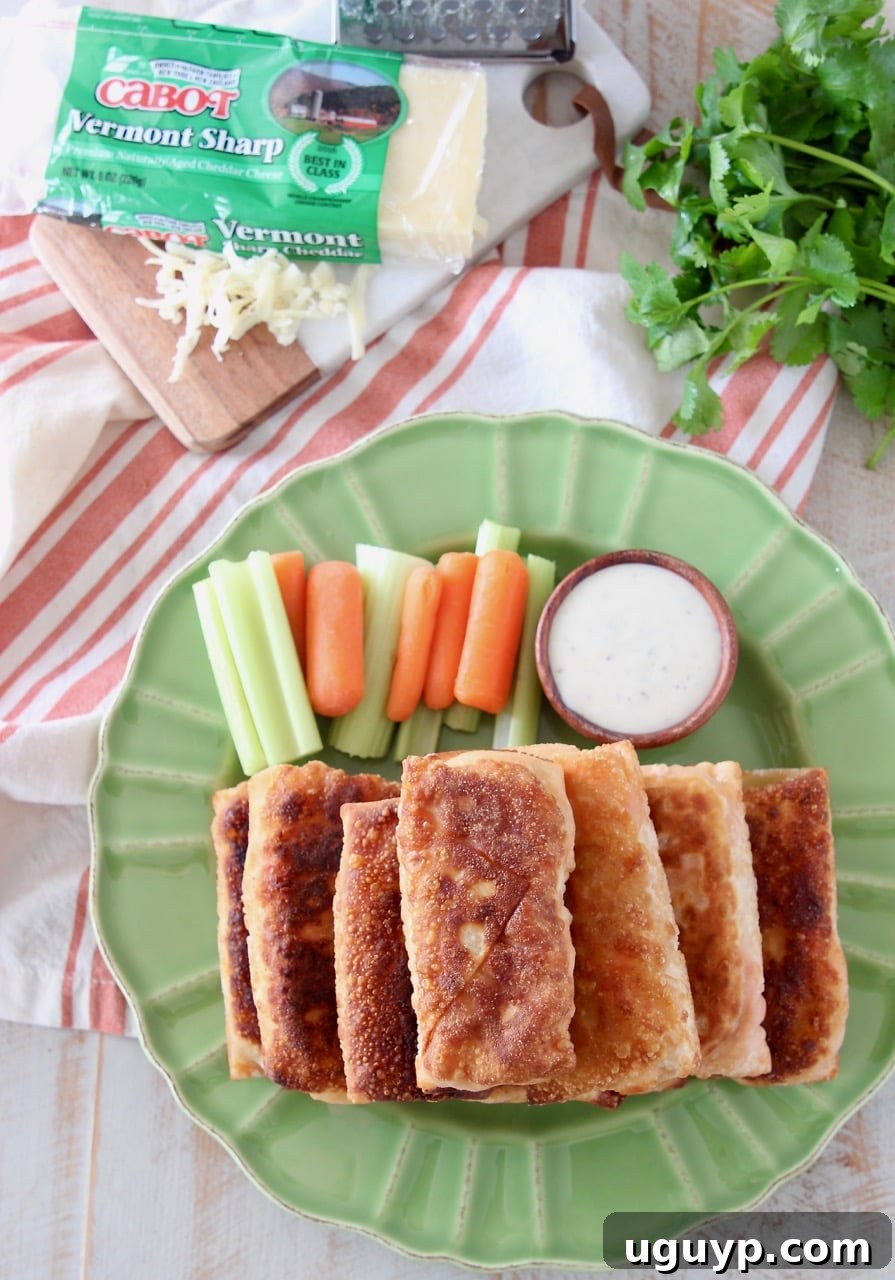 Overhead view of perfectly golden-brown buffalo chicken egg rolls stacked artfully on a green plate, accompanied by crisp carrot and celery sticks and a small wooden ramekin of creamy ranch dressing for dipping.