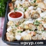 Cubed garlic bread covered in cheese on a baking sheet with a side of marinara sauce in a white bowl with a small spoon