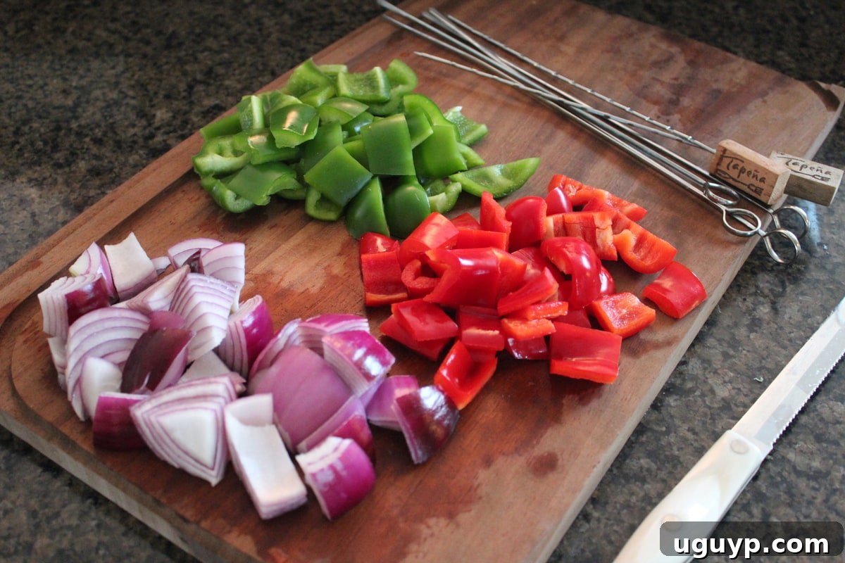 Diced red onions and colorful bell peppers neatly arranged on a wooden cutting board, with metal skewers ready for assembly.