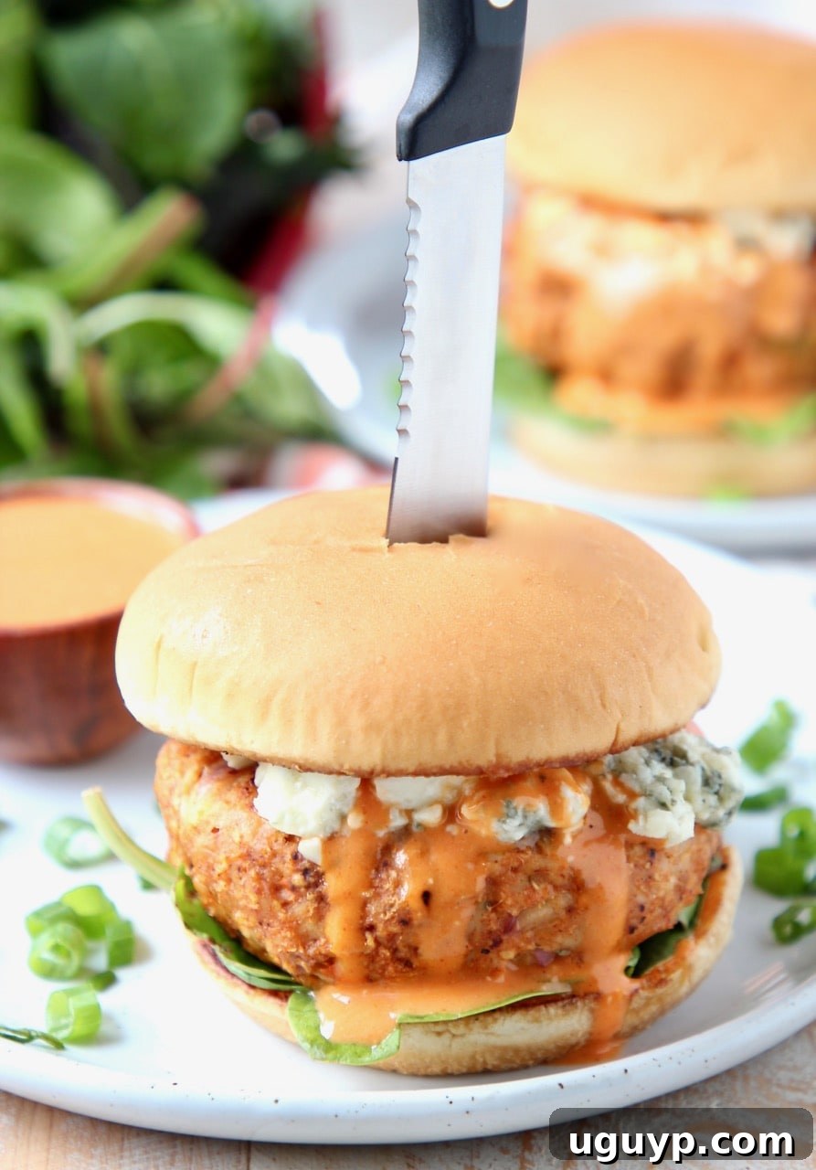 Buffalo chicken burger on plate, topped with blue cheese crumbles with knife in the middle of the burger