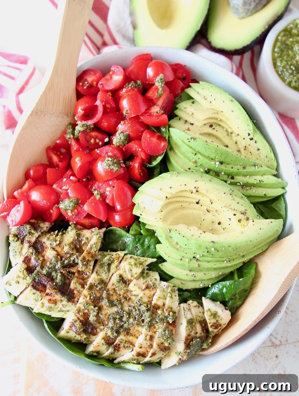 Sliced pesto chicken, sliced avocado, diced tomatoes and spinach in large bowl with wooden serving spoons