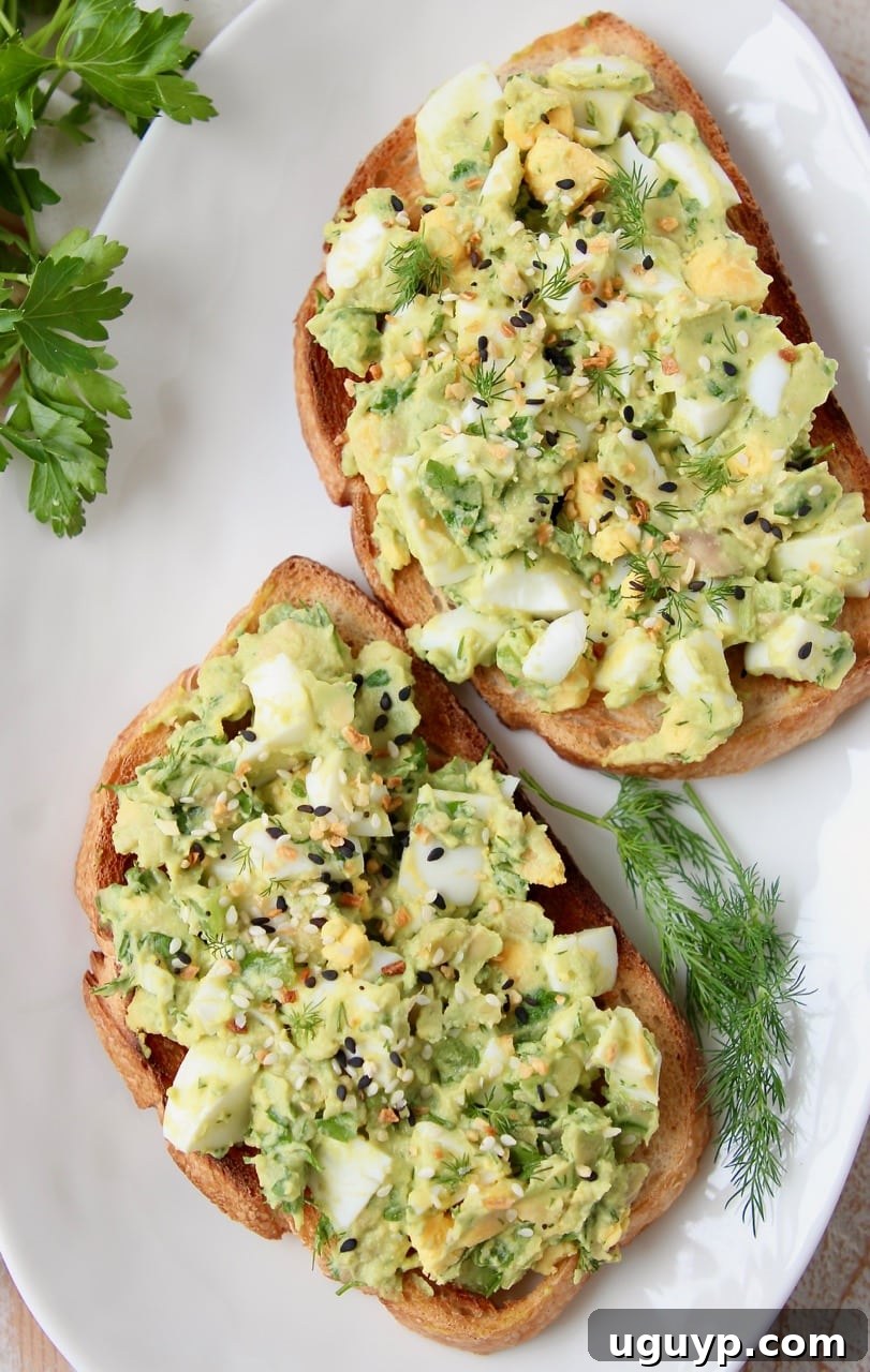 Two slices of sourdough toast topped with creamy avocado egg salad and fresh dill, presented on a white plate. Ideal for a healthy breakfast or lunch.
