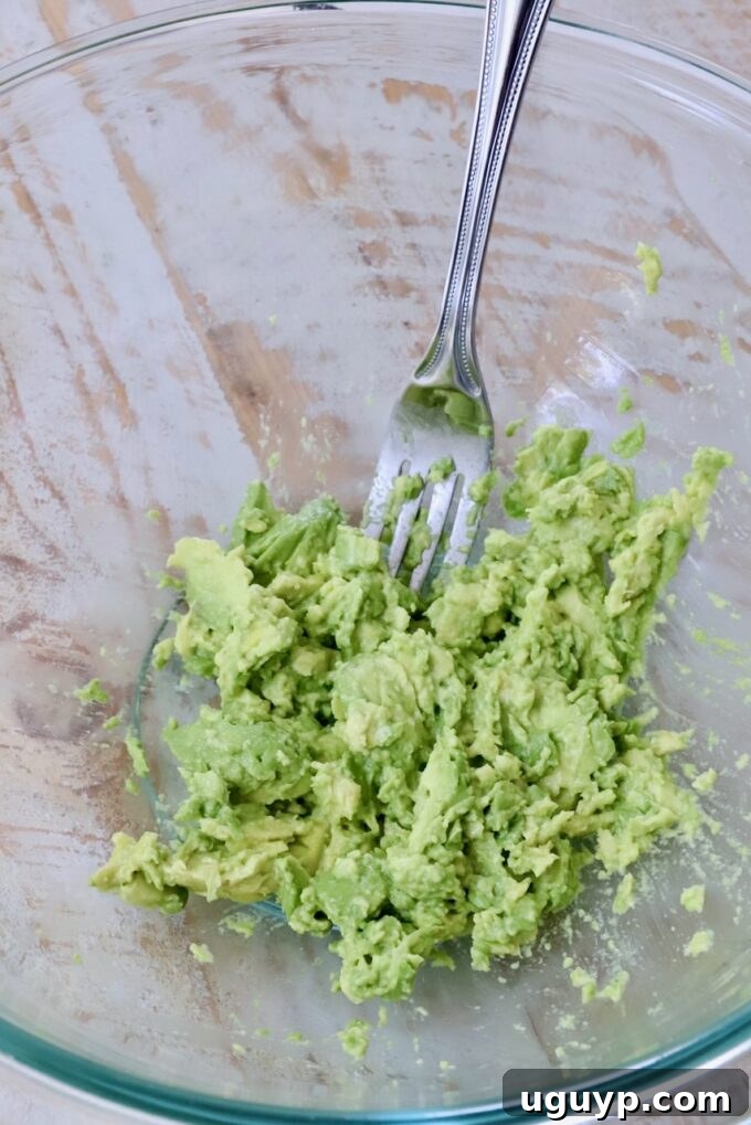 Roughly mashed avocado in a clear glass bowl with a fork, showing the ideal creamy yet chunky texture for avocado egg salad.