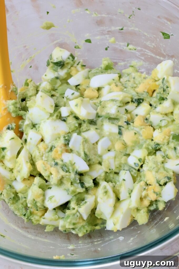 Creamy avocado egg salad mixed gently in a clear glass bowl with a yellow spatula, ready for serving.