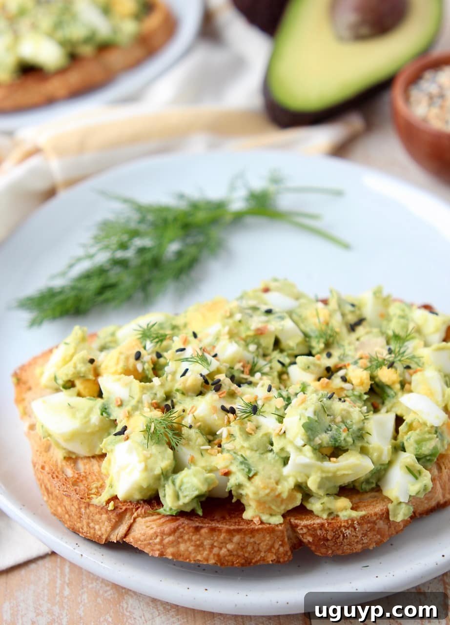 A slice of toasted sourdough bread topped with creamy avocado egg salad, garnished with fresh dill, on a simple white plate.