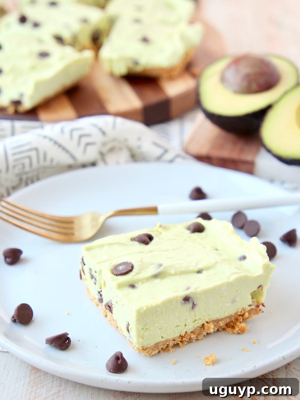Avocado cheesecake bar with graham cracker crust on white plate with gold fork and chocolate chips. A refreshing and easy summer dessert.