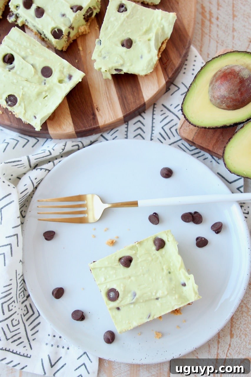 Overhead shot of avocado cheesecake bars on plate and cutting board with chocolate chips and halved avocado, showcasing the texture and vibrant color.