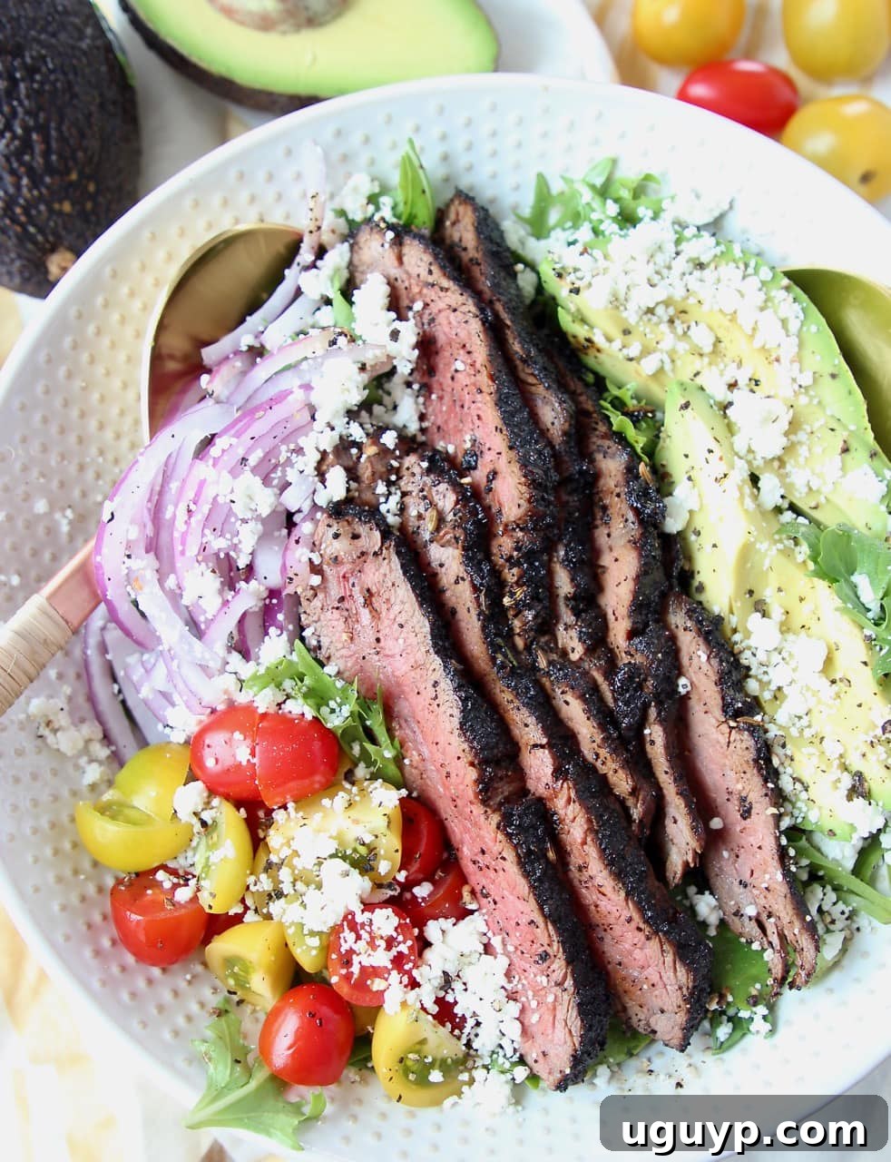 Grilled coffee-crusted flank steak salad in a large white bowl with avocado, tomatoes, and red onions, ready to be served.