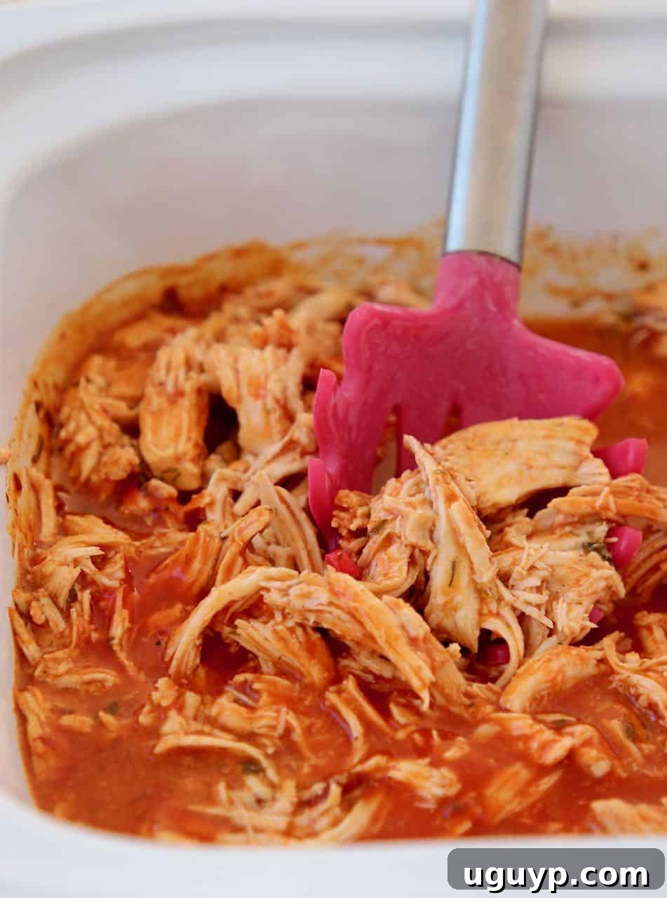 A close-up of tender, shredded buffalo chicken bubbling in a crock pot, with a red slotted serving spoon nestled in the delicious sauce.