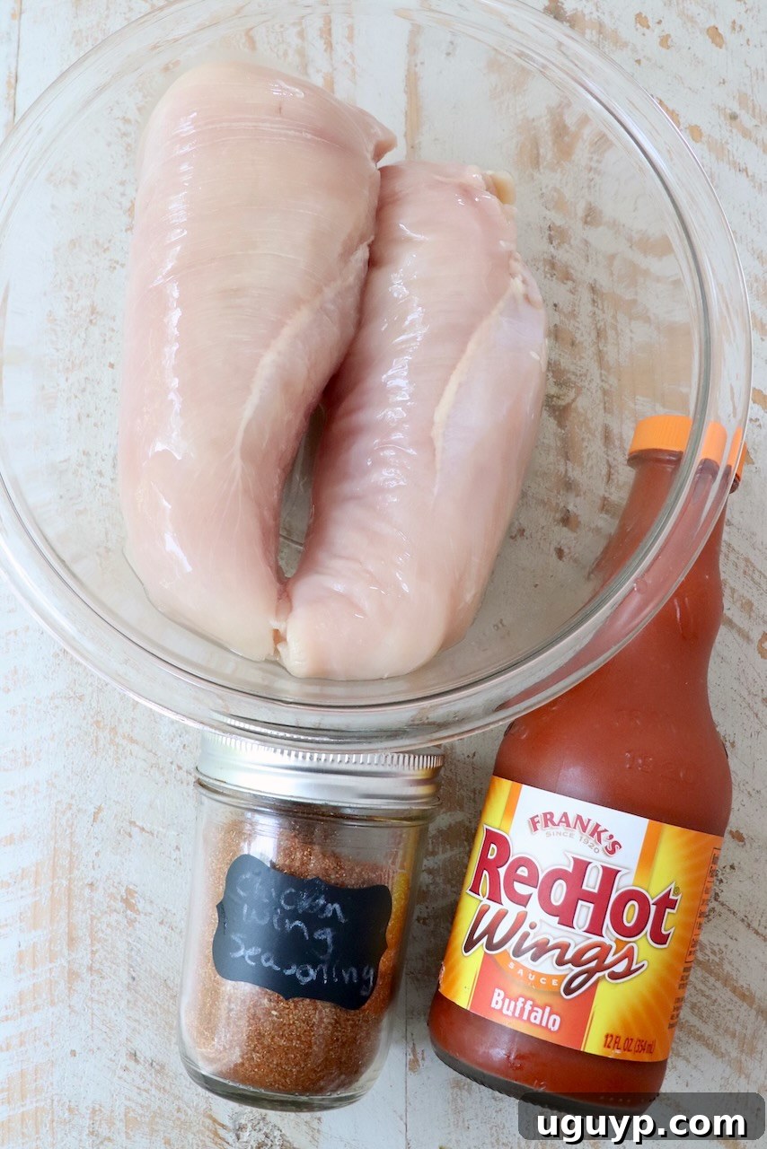 Selection of ingredients for slow cooker buffalo chicken on a rustic white wooden board, including chicken, buffalo sauce, and seasoning.