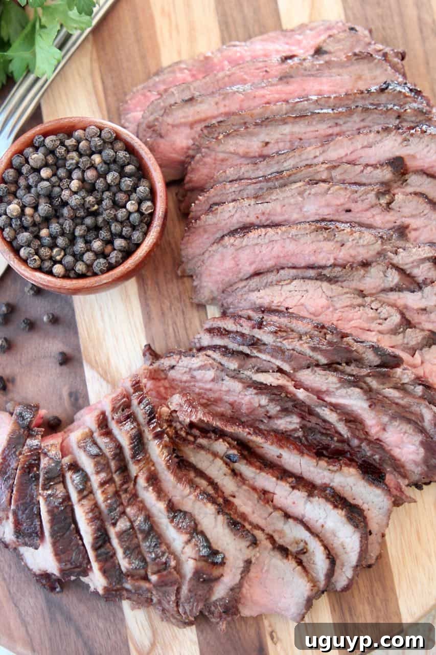 Sliced tri tip steak on wood cutting board with a bowl of black peppercorns