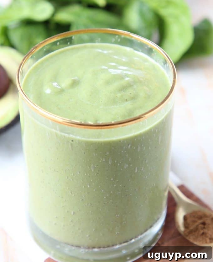 A refreshing Superfood Green Smoothie in a clear glass, positioned in front of fresh spinach leaves and a sliced avocado, emphasizing its natural and healthy ingredients.