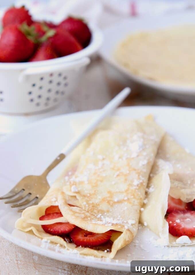 Beautifully folded French crepes, generously filled with Nutella and fresh sliced strawberries, elegantly arranged on a white plate with a fork, ready to be enjoyed as a sweet brunch or dessert.