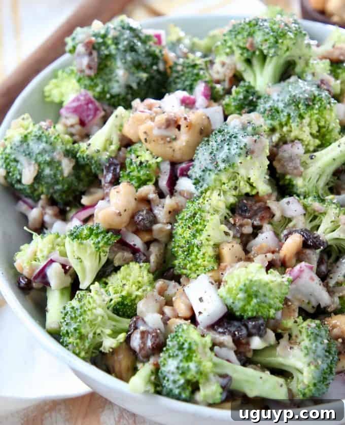 A large white bowl filled with a vibrant 10 Minute Broccoli Salad, featuring fresh broccoli florets, crispy bacon bits, sweet raisins, and crunchy cashews, all coated in a creamy dressing.