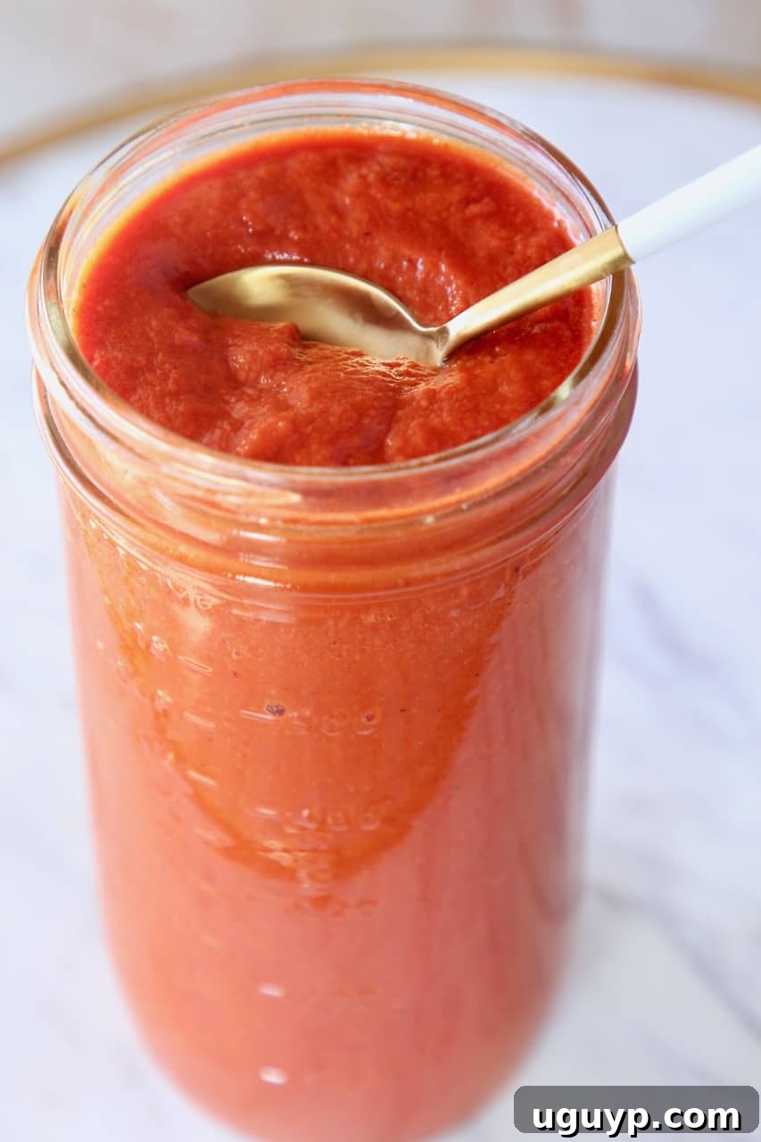 A spoon lifted from a mason jar of vibrant red enchilada sauce, showcasing its smooth texture and rich color.