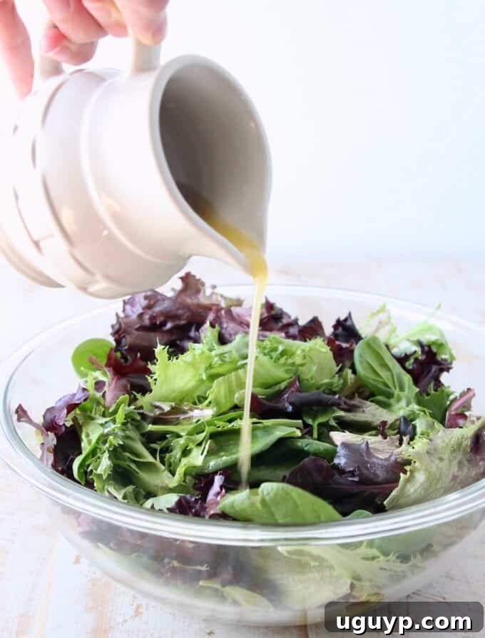 Small pitcher pouring vibrant homemade vinaigrette dressing over a fresh, colorful salad in a clear glass bowl, showcasing its appeal.